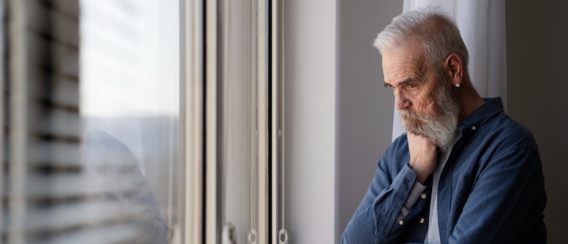 Hombre mayor mirando por la ventana pensativo y triste cabizbajo mano apoyada en la barbilla