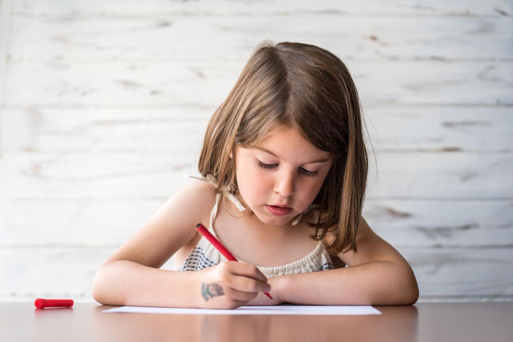 Niña concentrada escribiendo en un papel con un rotulador rojo.