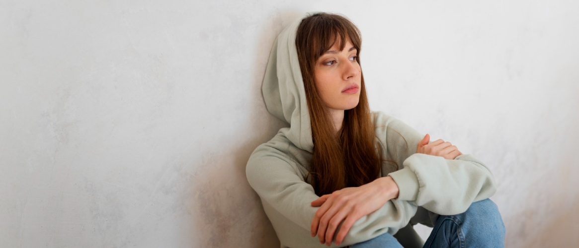 Chica joven apoyada en la pared pelo largo capucha manos cruzadas mirada perdida triste y abatida