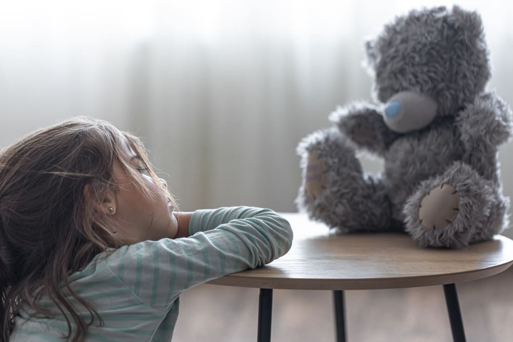 Niña triste apoyada en una mesita mira a su osito de peluche.