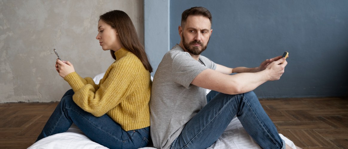 Pareja sentada apoyada espaldas mirando móviles él mirando a cámara