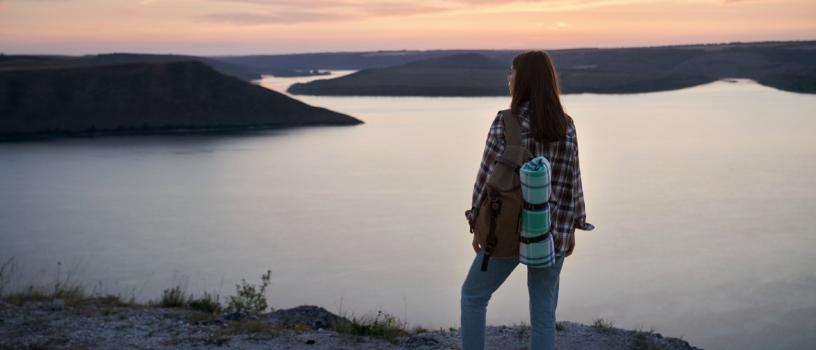 Mujer con mochila y esterilla en la espalda contemplando el paisaje en lo alto de una colina lago y montañas